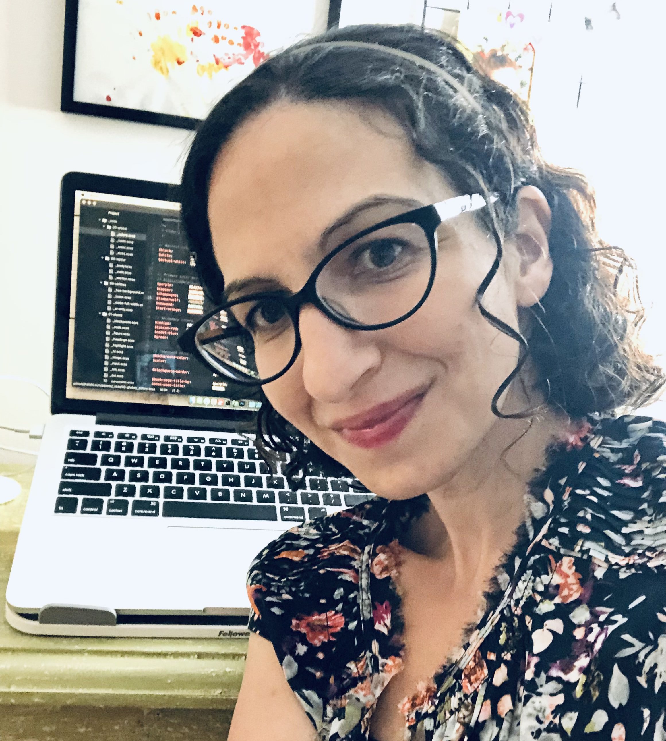 Joni, a white woman with dark curly hair and glasses, is sitting with her back to a desk. On the desk is an open laptop. The screen shows blurred out computer code.